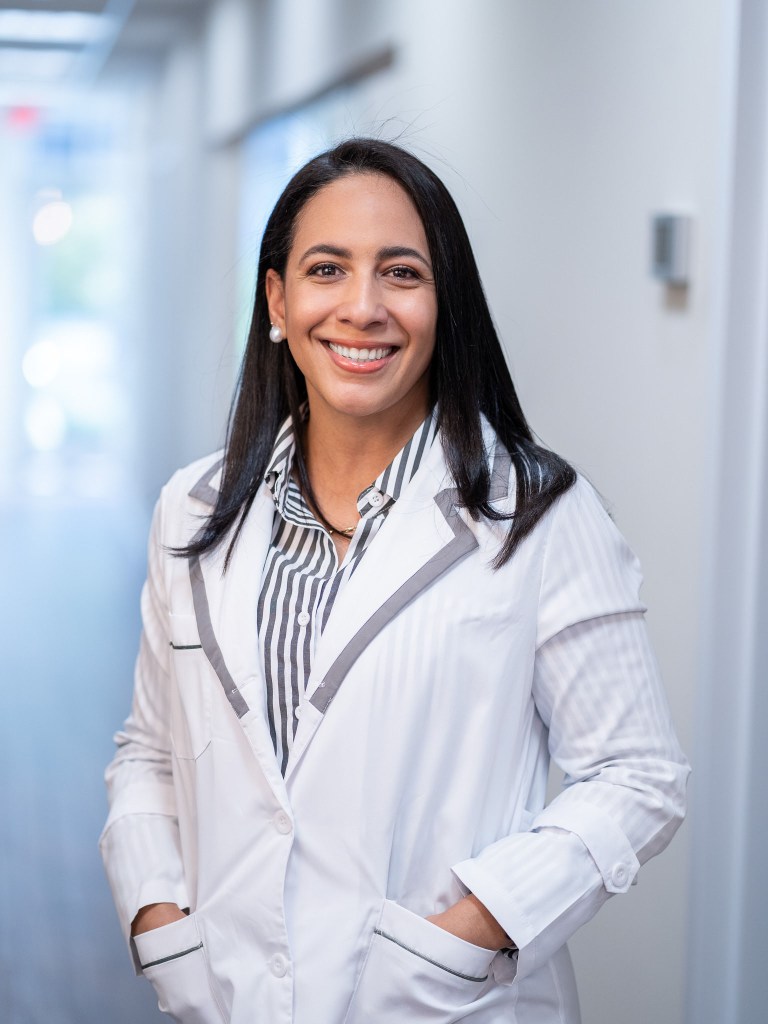 Professional headshot of Dr. Ivette Lora, DMD, smiling dentist with long dark hair wearing a white lab coat over a striped shirt in a modern office hallway.