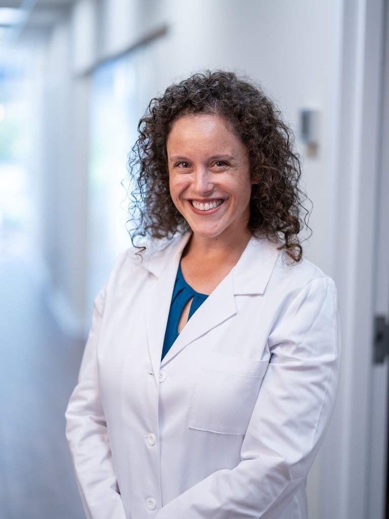Professional headshot of Dr. Adi Kedem, DMD, smiling dentist with curly hair wearing a white lab coat over a teal top in a modern office hallway.