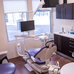 Dental operatory with modern equipment, purple patient chair, ceiling-mounted light and delivery system, computer workstation, and natural light from windows.