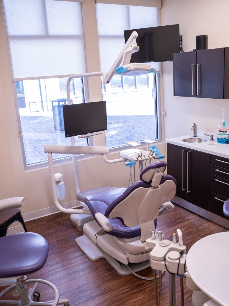 Dental operatory with modern equipment, purple patient chair, ceiling-mounted light and delivery system, computer workstation, and natural light from windows.