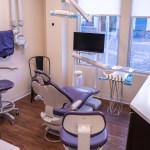 Dental operatory with modern equipment, purple patient chair, ceiling-mounted light and delivery system, computer workstation, and natural light from windows.