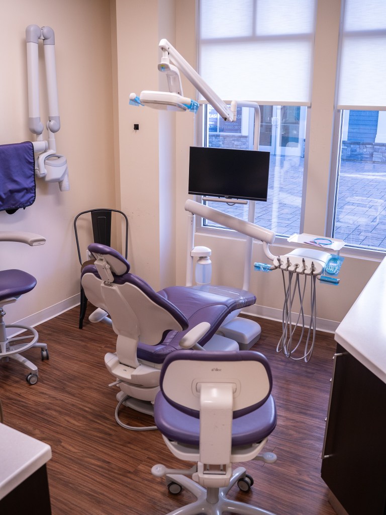 Dental operatory with modern equipment, purple patient chair, ceiling-mounted light and delivery system, computer workstation, and natural light from windows.